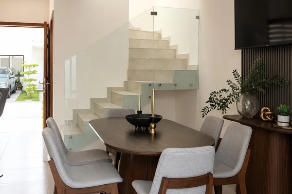 Modern dining room with round dark wood table, gray upholstered chairs, floating staircase with glass railing, and black ceiling fan.
