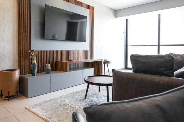 Modern living room with dark leather seating, wall-mounted TV with wood slat surround, white media console, round side table, and floor-to-ceiling windows.