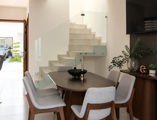 Comedor moderno con mesa redonda de madera oscura, sillas tapizadas en gris y, al fondo, una escalera flotante con barandilla de cristal.